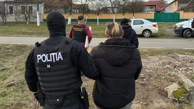 Un polițist cu mască neagră și vestă tactică stă lângă o persoană cu părul blond și un grup de oameni pe un teren de pietriș.
