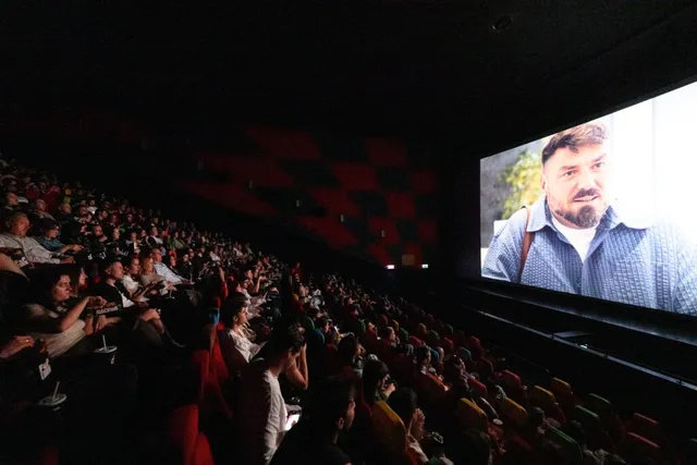 O sală de cinema întunecată cu spectatori așezați în rânduri de scaune, privind un ecran mare pe care apare un bărbat cu barbă.