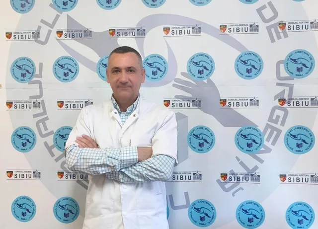 Un bărbat cu o halat albă de medic stă cu brațele încrucișate în fața unui perete decorat cu diverse sigilii și embleme circulare.
