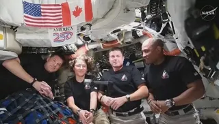 Patru persoane purtând tricouri negre stau într-un spațiu înghesuit plin de cabluri, echipamente și un steag american lângă unul canadian.