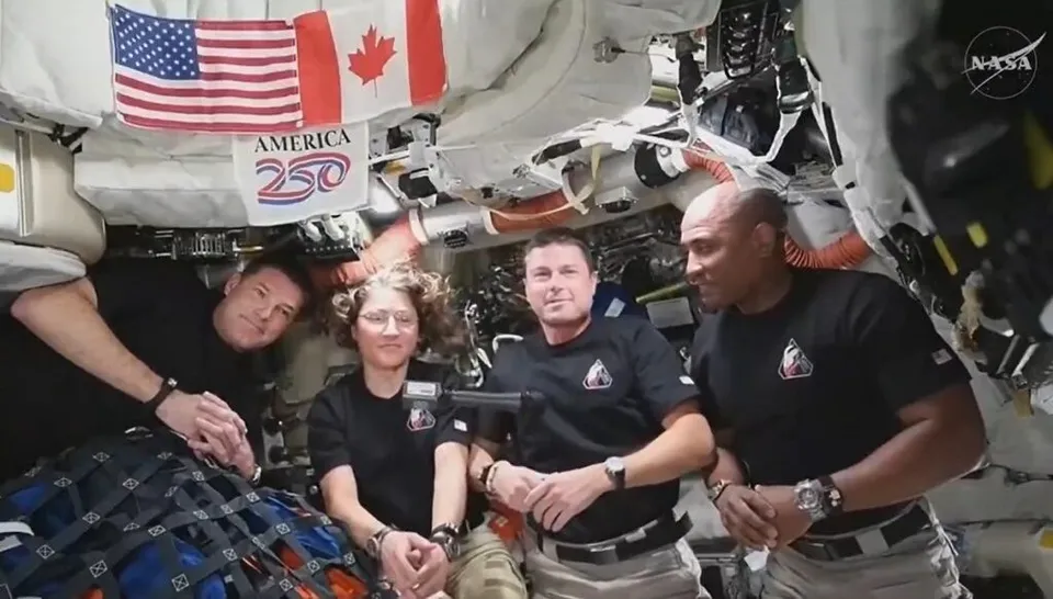 Patru persoane purtând tricouri negre stau într-un spațiu înghesuit plin de cabluri, echipamente și un steag american lângă unul canadian.