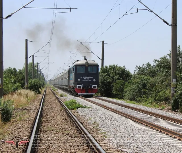 Un tren de pasageri rulează pe o cale ferată lângă linii electrice suspendate și vegetație verde sub un cer luminos.