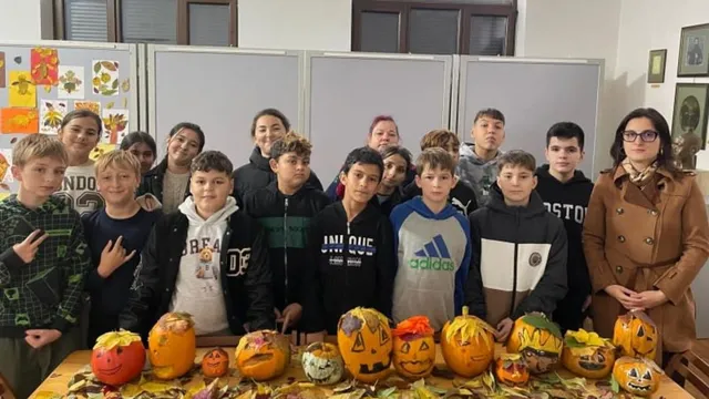 Un grup de copii și o femeie stau în spatele unei mese pe care sunt așezate diverse dovleci sculptați și pictați.