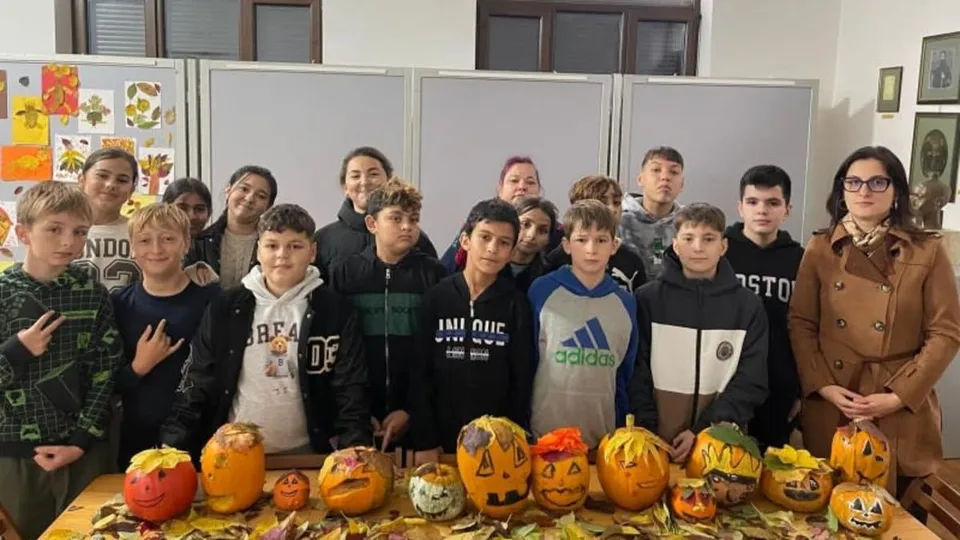 Un grup de copii și o femeie stau în spatele unei mese pe care sunt așezate diverse dovleci sculptați și pictați.