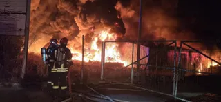 Doi pompieri în echipament de protecție stau lângă un gard metalic în fața unui incendiu cu flăcări mari și fum dens noaptea.