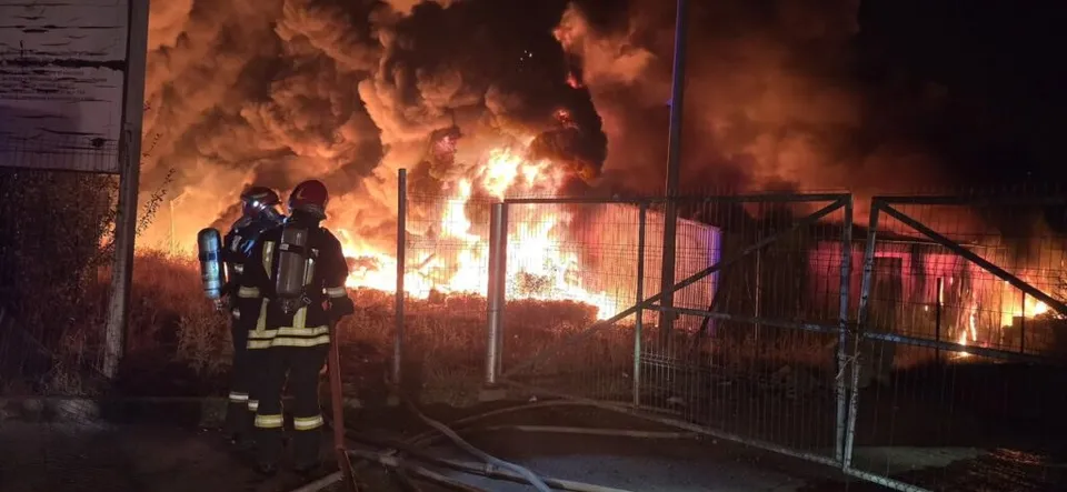 Doi pompieri în echipament de protecție stau lângă un gard metalic în fața unui incendiu cu flăcări mari și fum dens noaptea.