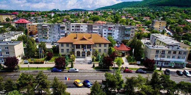 O vedere panoramică asupra unui oraș cu clădiri rezidențiale, copaci verzi și mașini parcate pe marginea unui drum în prim-plan.