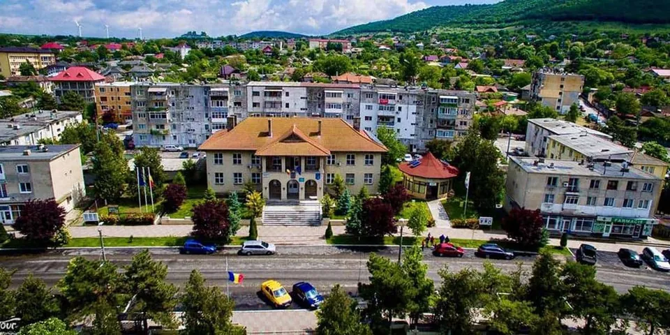O vedere panoramică asupra unui oraș cu clădiri rezidențiale, copaci verzi și mașini parcate pe marginea unui drum în prim-plan.