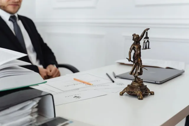 O statuetă de bronz reprezentând Justiția cu o balanță și o sabie stă pe un birou alb lângă un laptop și documente.
