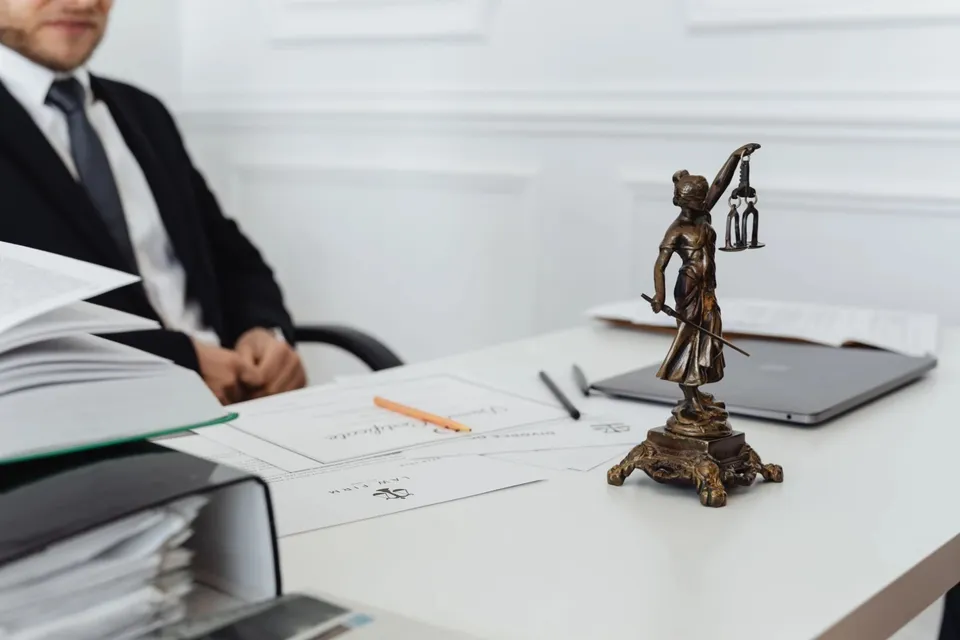 O statuetă de bronz reprezentând Justiția cu o balanță și o sabie stă pe un birou alb lângă un laptop și documente.