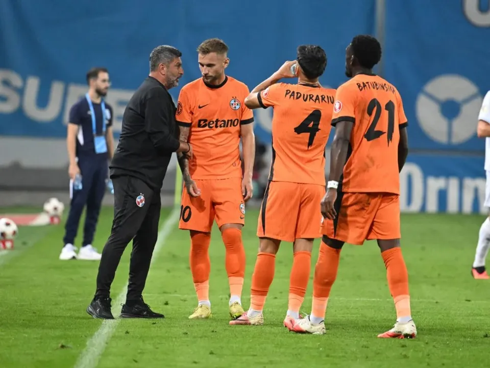 Fotbaliști în echipamente portocalii stau pe gazon lângă un bărbat în negru, în timp ce un alt bărbat privește din fundal.