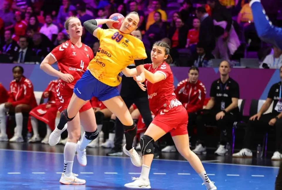 Două jucătoare în echipament roșu și una în echipament galben și albastru se află pe un teren de handbal în timpul unui meci.