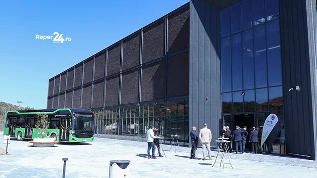 Oameni stau pe o esplanadă lângă un autobuz verde și negru în fața unei clădiri moderne cu fațadă de sticlă și panouri închise.