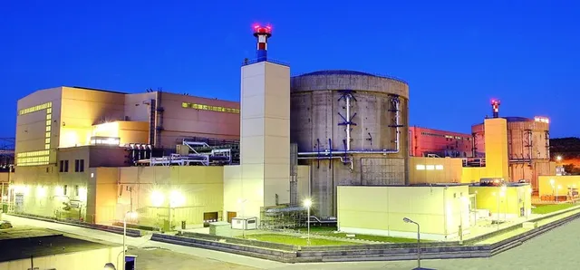 O centrală nucleară este iluminată noaptea, prezentând două containere cilindrice mari și diverse clădiri industriale sub un cer albastru închis.