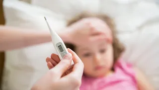 O persoană ține un termometru digital în prim-plan, în timp ce o altă mână atinge fruntea unui copil care stă în pat.