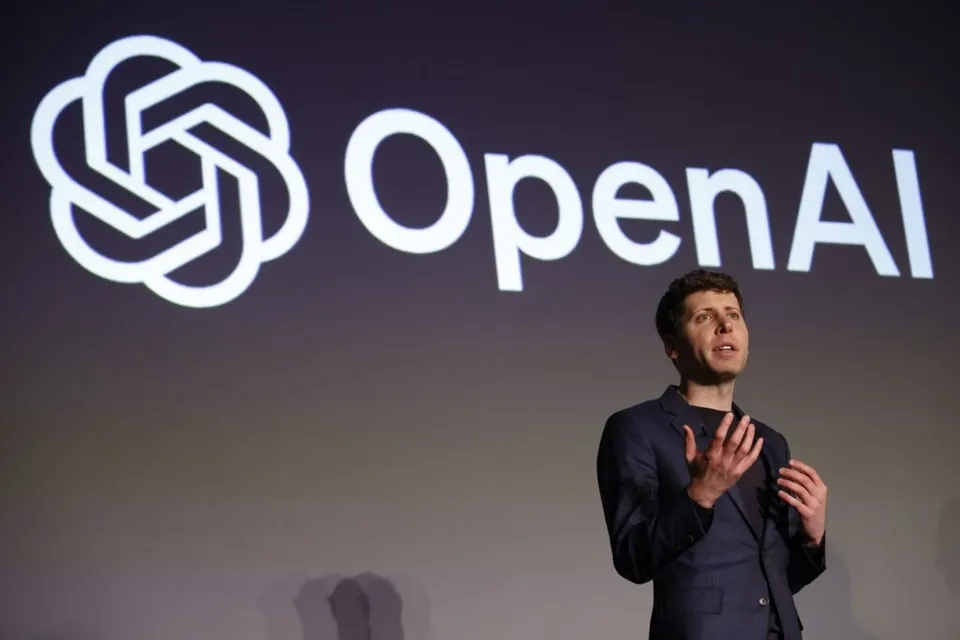 Un bărbat într-un costum închis stă în fața unui fundal gri pe care este proiectat un logo și textul OpenAI.