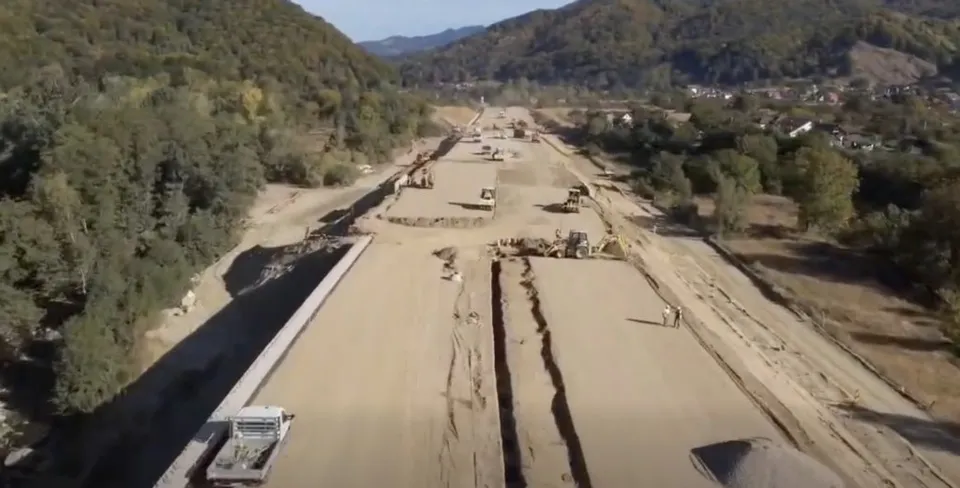 O vedere aeriană prezintă un șantier de drum cu teren nivelat, utilaje de construcții și dealuri împădurite în fundal.