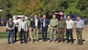Un grup de bărbați stă în rând pe un teren deschis, în fața unor copaci și vehicule.