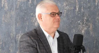 Un bărbat cu ochelari și sacou închis la culoare stă în fața unui microfon într-un cadru cu fundal gri texturat.