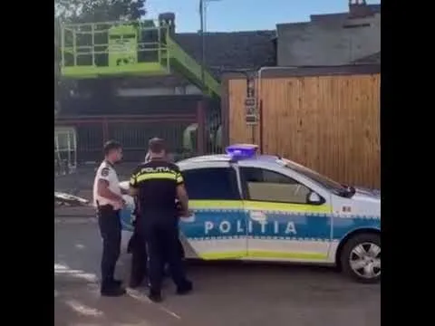 Un politist și un bărbat stau lângă o mașină de poliție albastră și albă cu lumini de semnalizare aprinse pe un drum asfaltat.