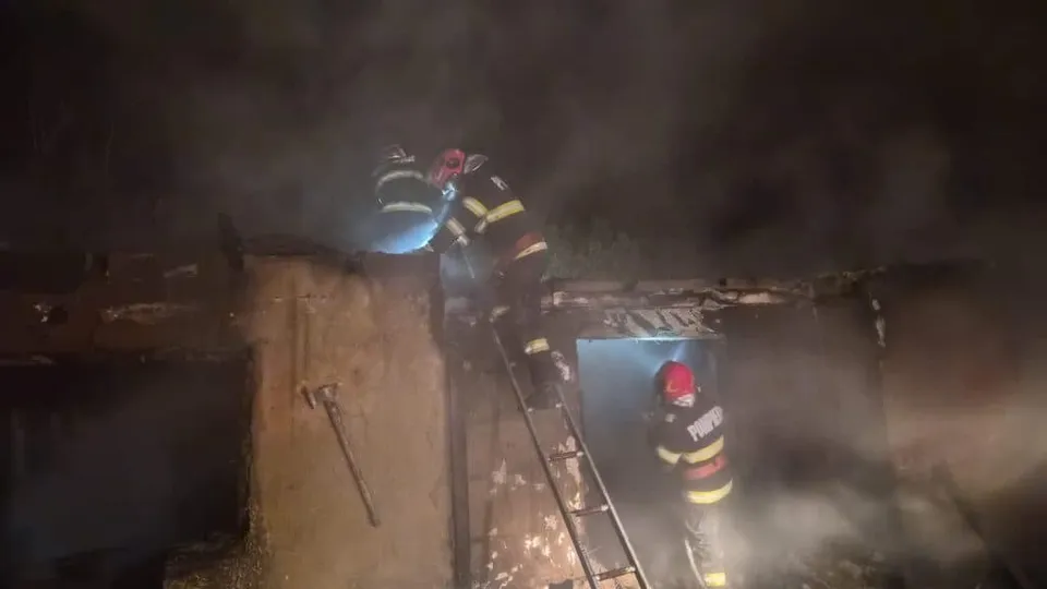 Pompieri cu echipament de protecție și cască stau pe o scară și lângă o clădire acoperită de fum noaptea.