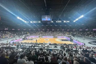 O mulțime de oameni stă pe scaune în tribunele unui stadion de baschet, privind spre terenul de joc iluminat.