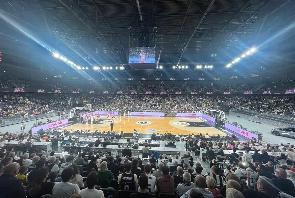 O mulțime de oameni stă pe scaune în tribunele unui stadion de baschet, privind spre terenul de joc iluminat.