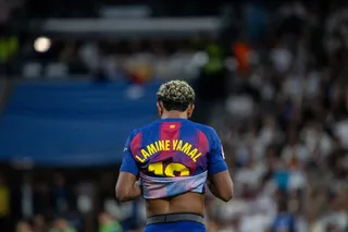 Un fotbalist cu părul blond și scurt, purtând un tricou albastru și roșu cu numele Lamine Yamal pe spate, stă cu spatele la cameră într-un stadion.