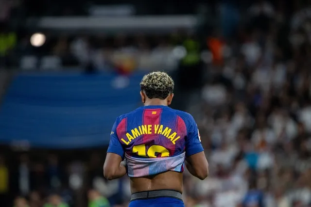 Un fotbalist cu părul blond și scurt, purtând un tricou albastru și roșu cu numele Lamine Yamal pe spate, stă cu spatele la cameră într-un stadion.