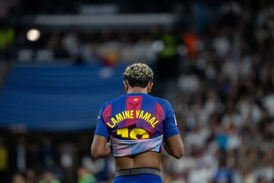 Un fotbalist cu părul blond și scurt, purtând un tricou albastru și roșu cu numele Lamine Yamal pe spate, stă cu spatele la cameră într-un stadion.