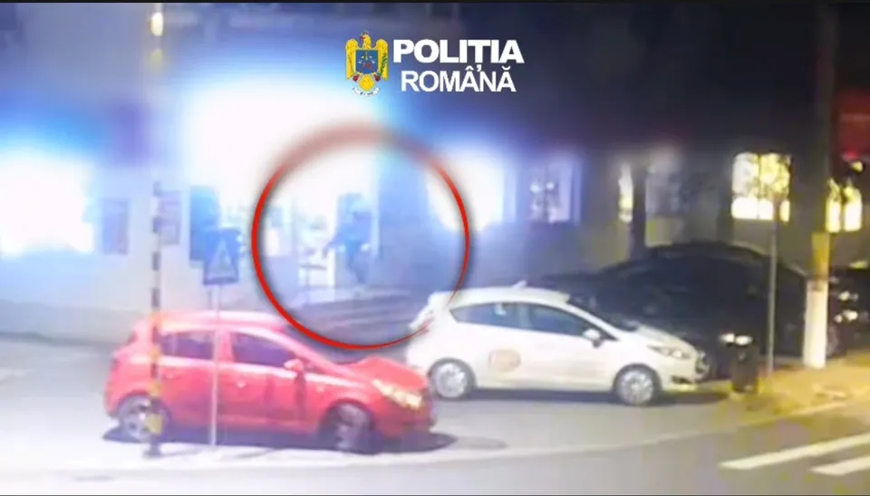 Două mașini, una roșie și una albă, sunt parcate pe o stradă lângă un clădire iluminată noaptea sub un logo al poliției.