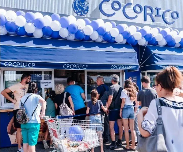 O mulțime de oameni stă la coadă în fața unui magazin cu un acoperiș de prețerie albastru decorat cu baloane albe și albastre.