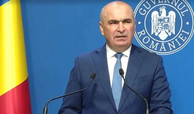 Un bărbat cu părul scurt și costum albastru, purtând o cămașă albă și o cravată albastră, stă în fața unui fundal albastru cu un emblemă circulară.