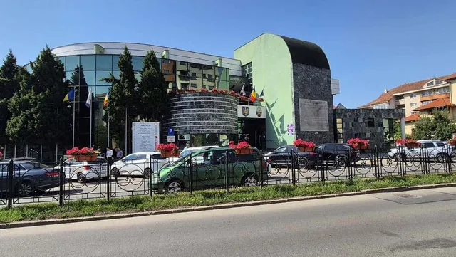 O clădire modernă cu fațadă de sticlă și un perete curbat de piatră se află în spatele unui gard metalic și a unor mașini parcate.