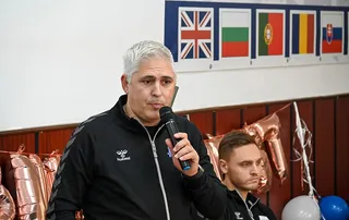 Un bărbat cu părul gri deschis ține un microfon negru și vorbește în fața unor steaguri și unor baloane metalice roz.