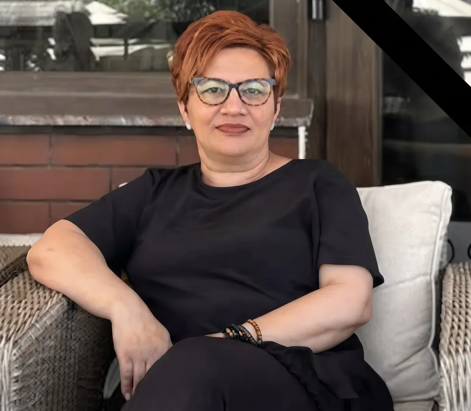 O femeie cu părul scurt roșu și ochelari cu rame negre poartă un tricou negru și stă pe o canapea cu perne albe.