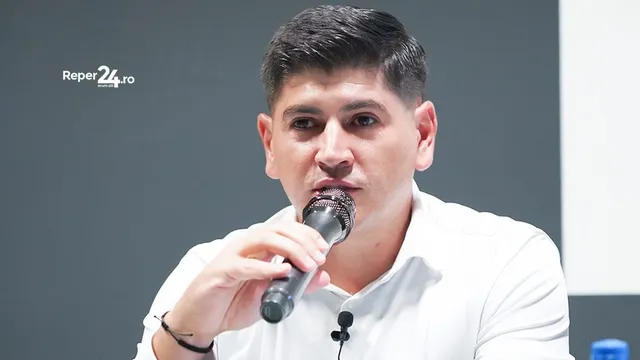 Un bărbat cu cămașă albă ține un microfon negru în mână și pare să vorbească în fața unui fundal gri.