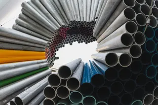 Multe țevi metalice de diferite dimensiuni și culori sunt stivuite într-un depozit sub un acoperiș.