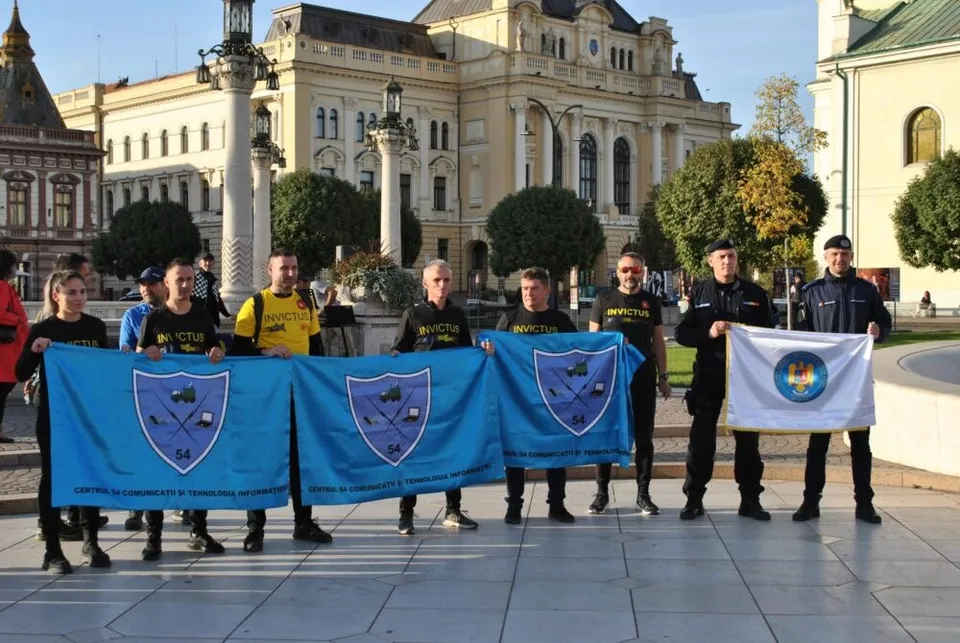 Un grup de bărbați stă în rând pe o piață pavată, ținând patru steaguri albastre și unul alb cu embleme colorate.