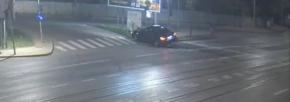 Un autoturism de culoare închisă este poziționat pe marginea unei străzi lângă un stâlp, sub lumina nocturnă.