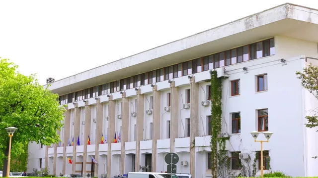 O clădire albă cu ferestre dreptunghiulare și vegetație verde pe margini este situată sub un cer deschis.