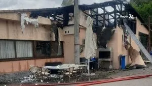 O clădire cu fațada afectată de arsuri și acoperișul distrus prezintă resturi de materiale și mese de exterior pe un teren de pietriș.