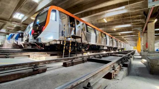 Un tren de metrou alb cu detalii portocalii este poziționat pe șine într-un depozit subteran cu tavan de beton și iluminat artificial.
