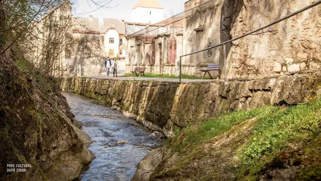Un râu curge printre maluri stâncoase, lângă un drum pavat cu bănci și clădiri vechi de piatră în fundal.