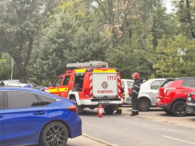 Un pompier cu cască roșie stă lângă un autospecial de pompieri alb și roșu parcat pe un drum lângă mașini colorate.