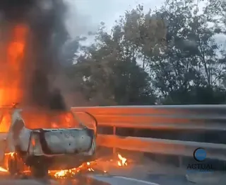 Un autoturism de culoare deschisă este cuprins de flăcări pe marginea unui drum lângă o barieră metalică de siguranță și copaci.