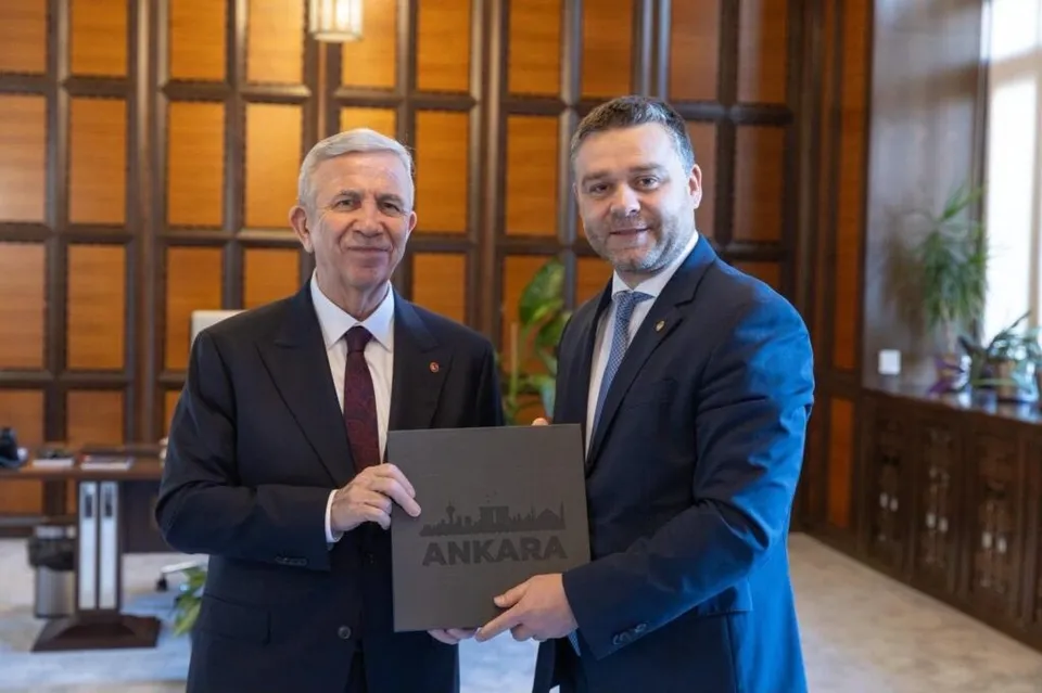 Doi bărbați în costume purtând cravate stau în picioare într-o încăpere cu panouri de lemn și țin împreună un dosar negru cu textul ANKARA.