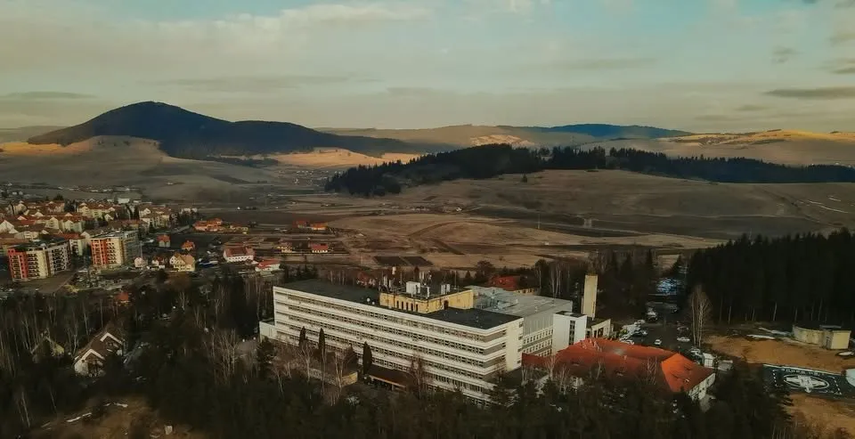 O vedere aeriană a unui oraș cu clădiri rezidențiale, dealuri împădurite și un complex de clădiri albe în prim-plan sub un cer înnorat.
