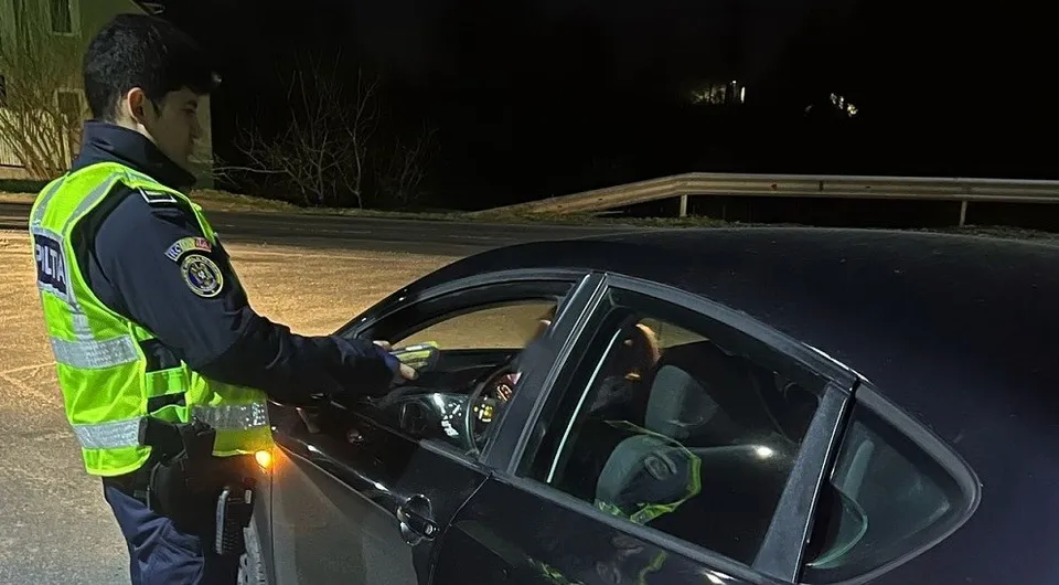 Un polițist în uniformă cu vestă reflectorizantă stă lângă fereastra deschisă a unei mașini negre noaptea.