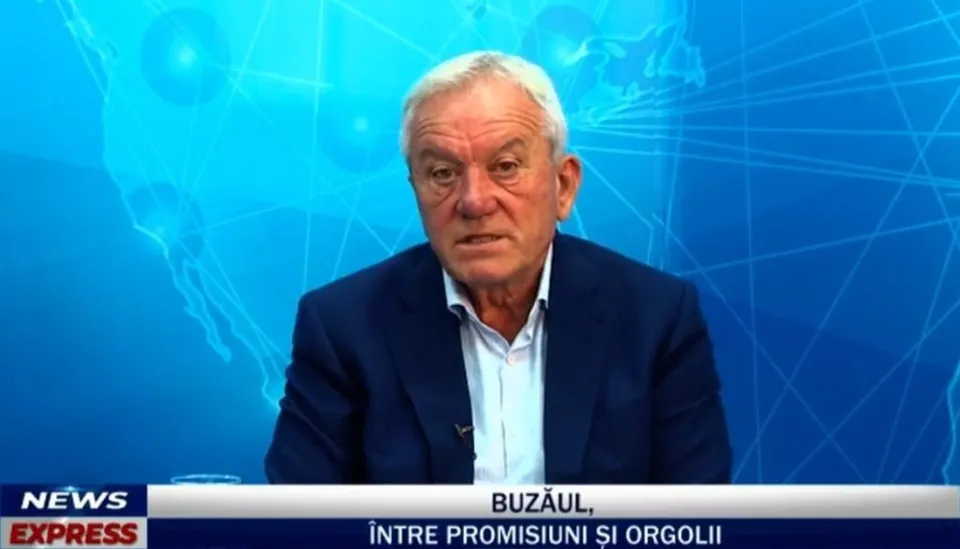 Un bărbat cu părul alb, purtând sacou închis și cămașă deschisă la guler, este poziționat în fața unui fundal albastru cu grafică digitală.